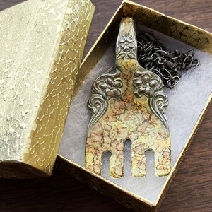 May include: A silver fork necklace with a decorative floral design and a marbled finish. The fork is hanging in a small, gold gift box with a black chain.