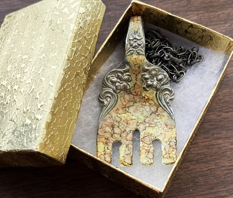 May include: A silver fork necklace with a decorative floral design and a marbled finish. The fork is hanging in a small, gold gift box with a black chain.