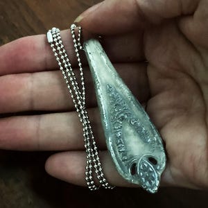 May include: A silver pendant necklace with a detailed, ornate design. The pendant is shaped like a long, narrow teardrop and has a textured surface. The necklace is made of a silver ball chain.