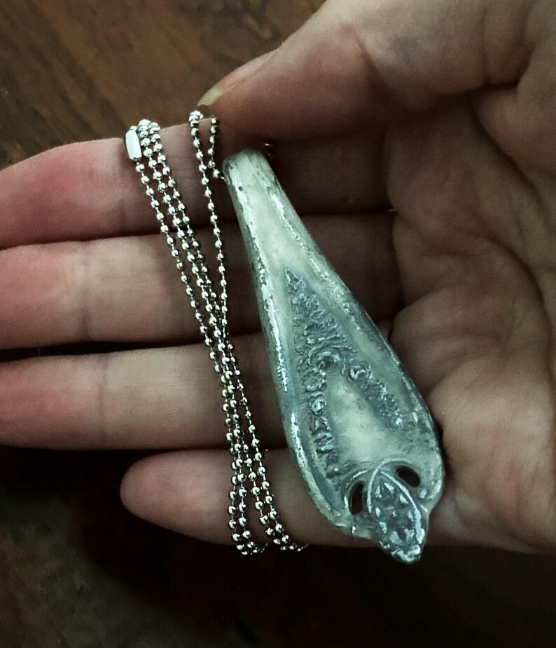 May include: A silver pendant necklace with a detailed, ornate design. The pendant is shaped like a long, narrow teardrop and has a textured surface. The necklace is made of a silver ball chain.