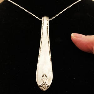 Puede incluir: Un collar con un colgante de cuchara de plata con un dise&ntilde;o floral decorativo en la parte inferior. El colgante est&aacute; unido a una cadena de plata.