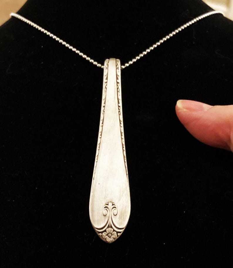 Puede incluir: Un collar con un colgante de cuchara de plata con un dise&ntilde;o floral decorativo en la parte inferior. El colgante est&aacute; unido a una cadena de plata.