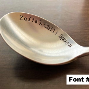 May include: A silver spoon with the text "Zofia's Chili Spoon" engraved on the bowl. The spoon is on a brown wooden surface. Font #2.