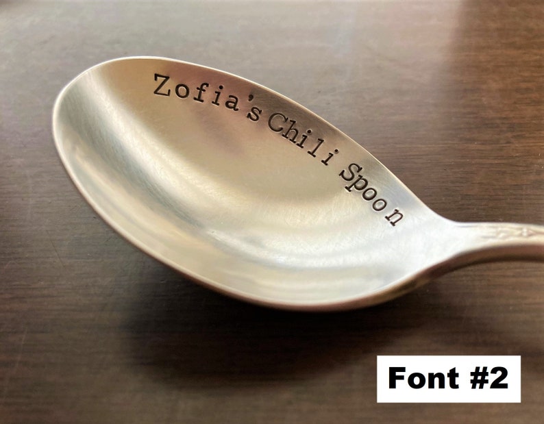 May include: A silver spoon with the text "Zofia's Chili Spoon" engraved on the bowl. The spoon is on a brown wooden surface. Font #2.