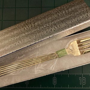 May include: A gold-colored fork with a decorative handle is displayed in a box. The fork is made of metal and has a polished finish. The box is lined with white paper and has a silver-colored exterior.
