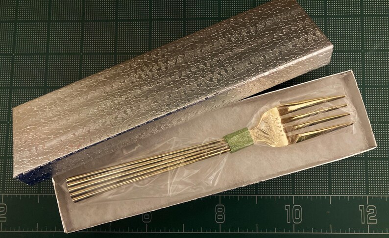 May include: A gold-colored fork with a decorative handle is displayed in a box. The fork is made of metal and has a polished finish. The box is lined with white paper and has a silver-colored exterior.