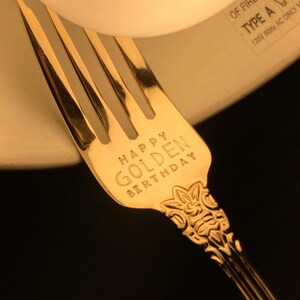 May include: A gold-colored fork with the words "HAPPY GOLDEN BIRTHDAY" engraved on the handle. The fork has a decorative floral design at the base of the handle and is set against a blurred background.