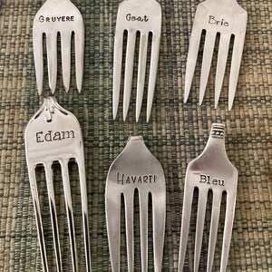 May include: Six silver forks with different cheese names engraved on them: Gruyere, Goat, Brie, Edam, Havarti, and Bleu.