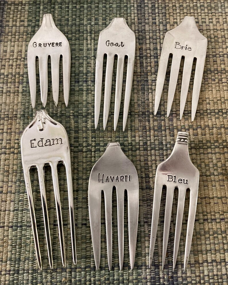 May include: Six silver forks with different cheese names engraved on them: Gruyere, Goat, Brie, Edam, Havarti, and Bleu.