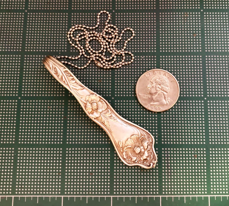 May include: A silver spoon necklace with a floral design. The spoon is attached to a silver chain. The spoon is about the size of a quarter.