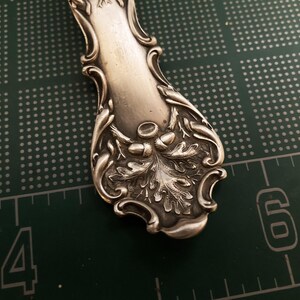 May include: Close-up of the ornate handle of a silver spoon, featuring an intricate design of oak leaves and acorns.