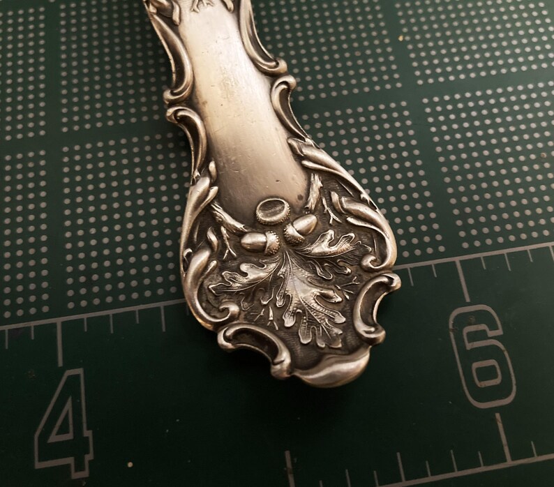 May include: Close-up of the ornate handle of a silver spoon, featuring an intricate design of oak leaves and acorns.