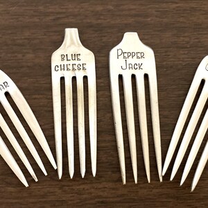 May include: Four silver forks with different cheese names engraved on the handles: Cheddar, Blue Cheese, Pepper Jack, and Gouda.