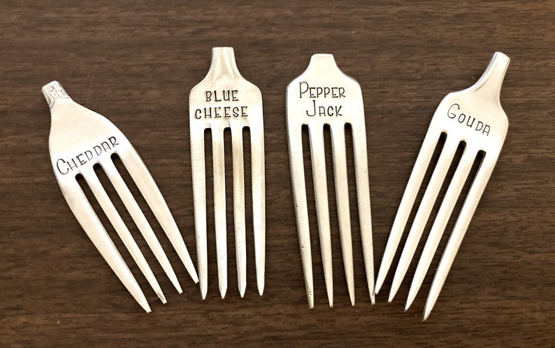 May include: Four silver forks with different cheese names engraved on the handles: Cheddar, Blue Cheese, Pepper Jack, and Gouda.