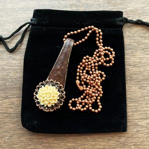 May include: A necklace with a copper chain and a pendant featuring a yellow flower with a brown and black metal design. The necklace is in a black velvet pouch.