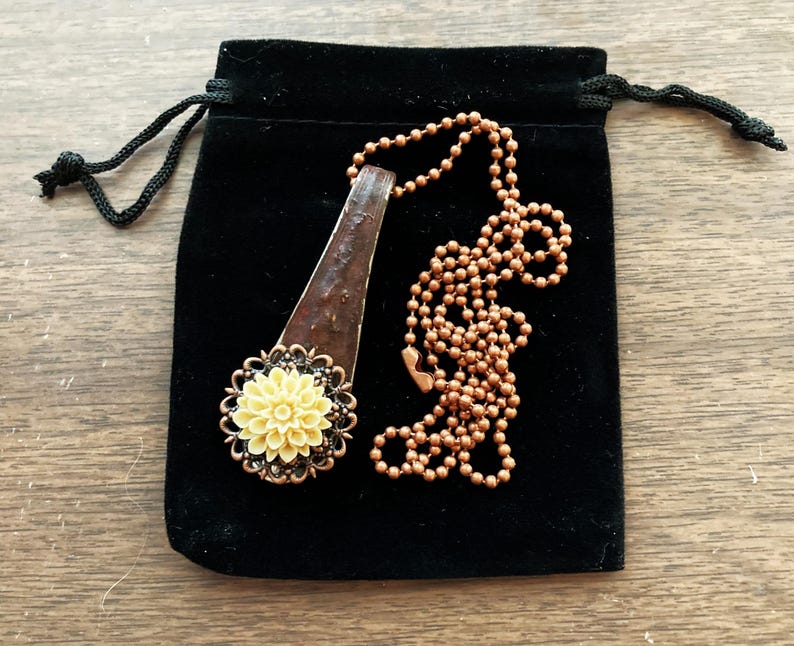 May include: A necklace with a copper chain and a pendant featuring a yellow flower with a brown and black metal design. The necklace is in a black velvet pouch.