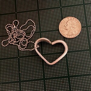 Puede incluir: Un collar con un colgante en forma de coraz&oacute;n de plata y una cadena de plata. El colgante est&aacute; abierto por un lado y la cadena est&aacute; unida por el otro lado. El collar est&aacute; sobre una superficie verde.