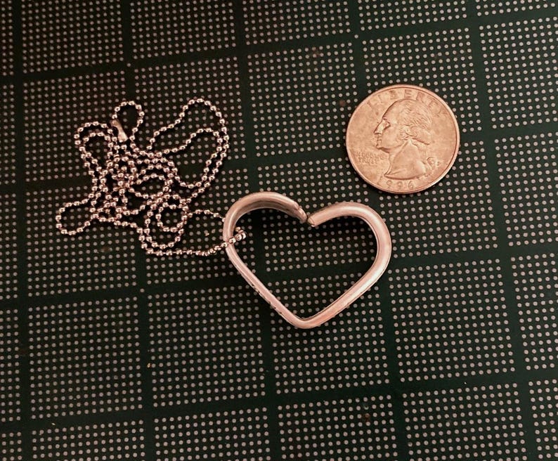 Puede incluir: Un collar con un colgante en forma de coraz&oacute;n de plata y una cadena de plata. El colgante est&aacute; abierto por un lado y la cadena est&aacute; unida por el otro lado. El collar est&aacute; sobre una superficie verde.
