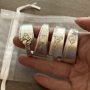 May include: Five silver spoon rings with decorative floral designs. The rings are stacked on top of each other and are displayed on a hand.