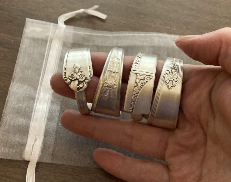 May include: Five silver spoon rings with decorative floral designs. The rings are stacked on top of each other and are displayed on a hand.