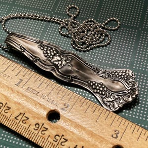 May include: Silver necklace with a vintage spoon pendant featuring a grapevine design. The spoon is about 2 inches long and is attached to a silver chain.