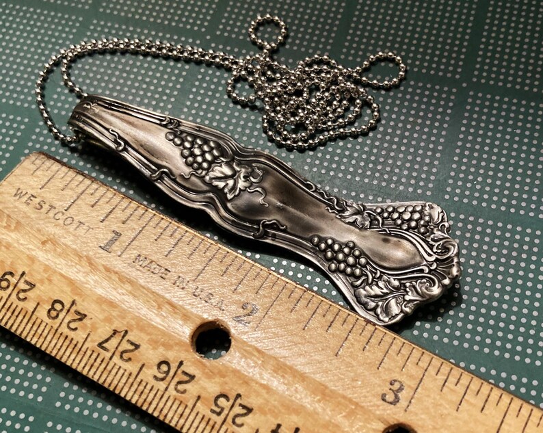 May include: Silver necklace with a vintage spoon pendant featuring a grapevine design. The spoon is about 2 inches long and is attached to a silver chain.