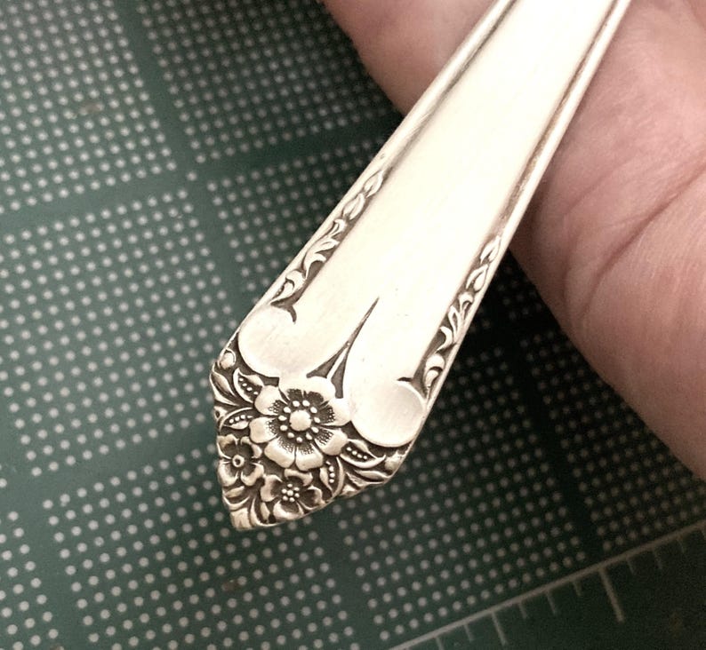 May include: Close-up of a silver-colored utensil, possibly a spoon or fork, with intricate floral and leaf patterns. The handle features a detailed flower design at the base, with delicate curves and embellishments along the edges. The utensil is held against a green surface.