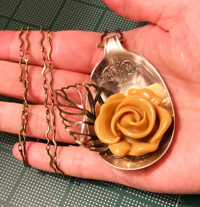 May include: A gold chain necklace with a vintage spoon pendant featuring a brown rose and a leaf design.