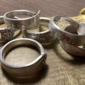 May include: Five silver spoon rings with intricate floral designs. The rings are stacked on a gold fabric surface.