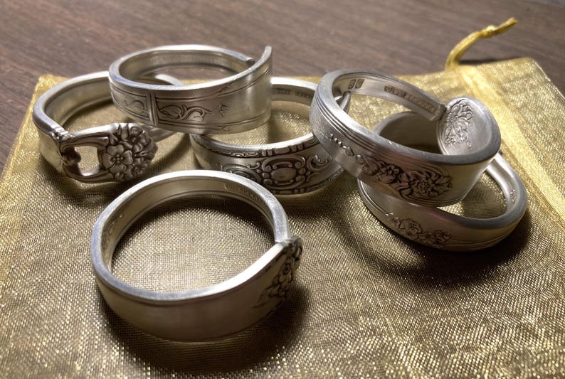 May include: Five silver spoon rings with intricate floral designs. The rings are stacked on a gold fabric surface.