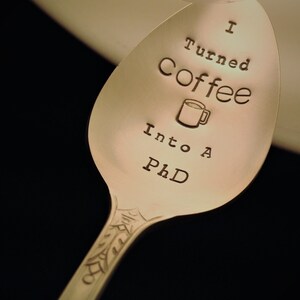 Pu&ograve; includere: Cucchiaio d'argento con la scritta incisa "I Turned Coffee Into A PhD". Un disegno di tazza di caff&egrave; &egrave; presente. Il manico del cucchiaio presenta un motivo decorativo.