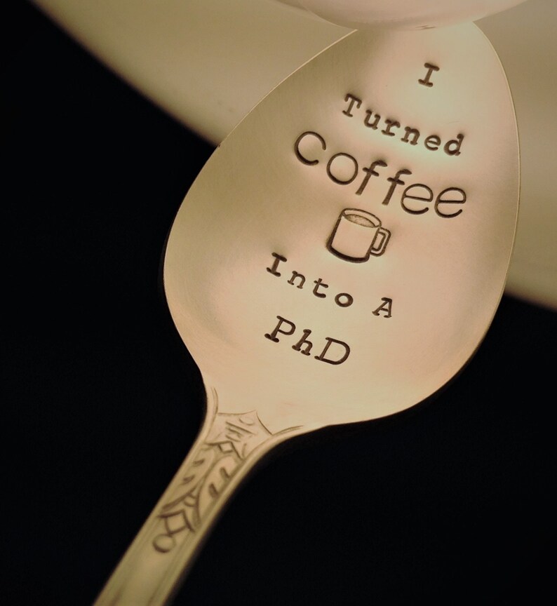 Pu&ograve; includere: Cucchiaio d'argento con la scritta incisa "I Turned Coffee Into A PhD". Un disegno di tazza di caff&egrave; &egrave; presente. Il manico del cucchiaio presenta un motivo decorativo.