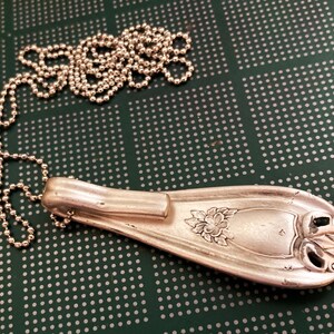 May include: A silver spoon necklace with a floral design and a ball chain. The spoon is bent into a pendant shape and has a decorative handle.