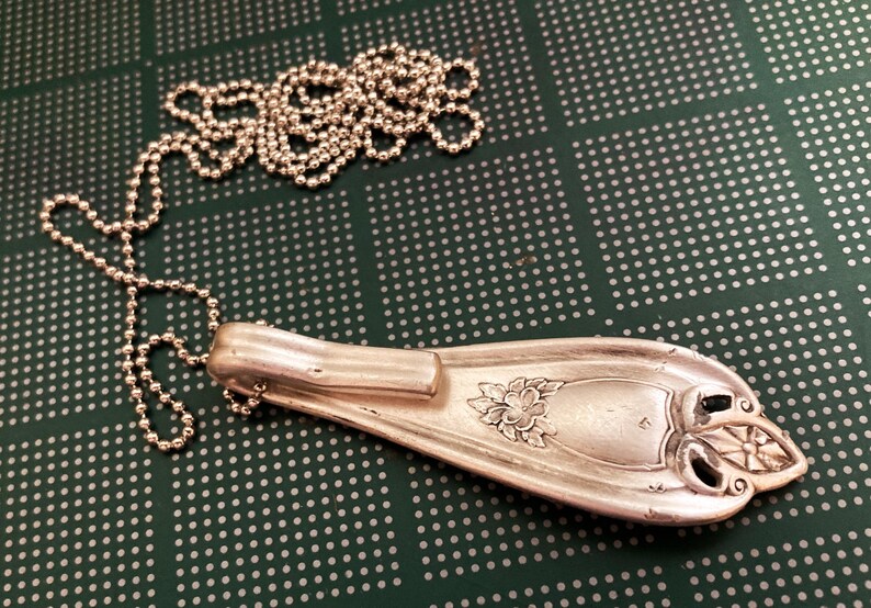 May include: A silver spoon necklace with a floral design and a ball chain. The spoon is bent into a pendant shape and has a decorative handle.