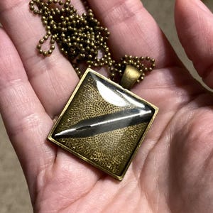 K&ouml;nnte beinhalten: Eine goldene Kettenkette mit einem quadratischen Anh&auml;nger, der einen silbernen Federkiel in einem quadratischen, klaren Harz enth&auml;lt.