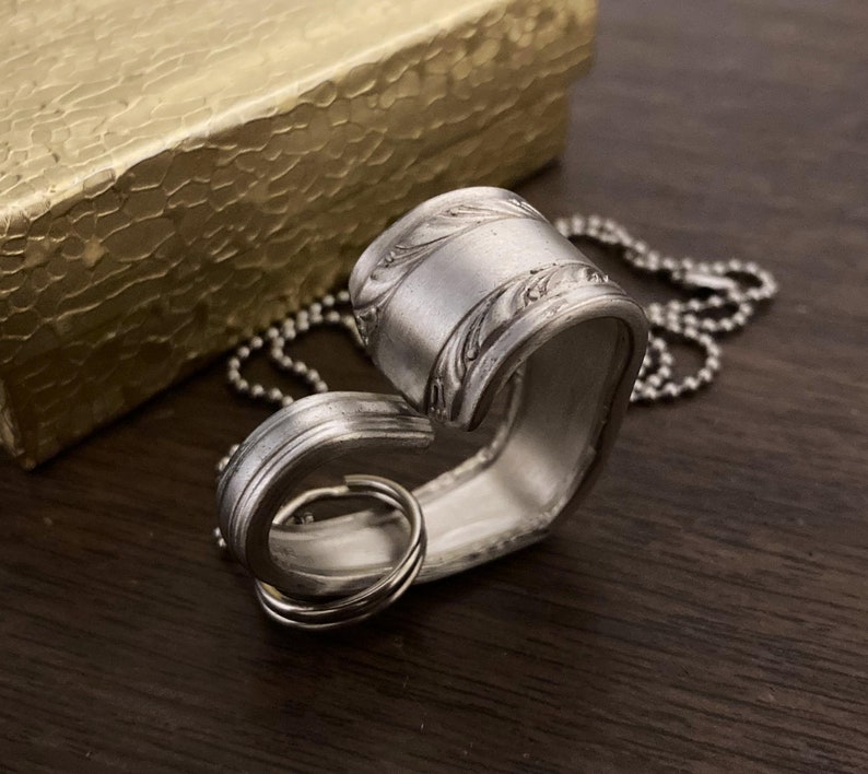 May include: A silver heart-shaped pendant necklace made from a repurposed spoon. The pendant has a detailed floral design and is attached to a silver ball chain.