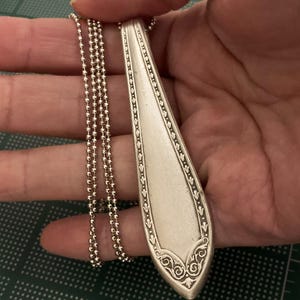 May include: A silver spoon pendant necklace with a decorative design on the handle. The necklace is made with a silver ball chain.