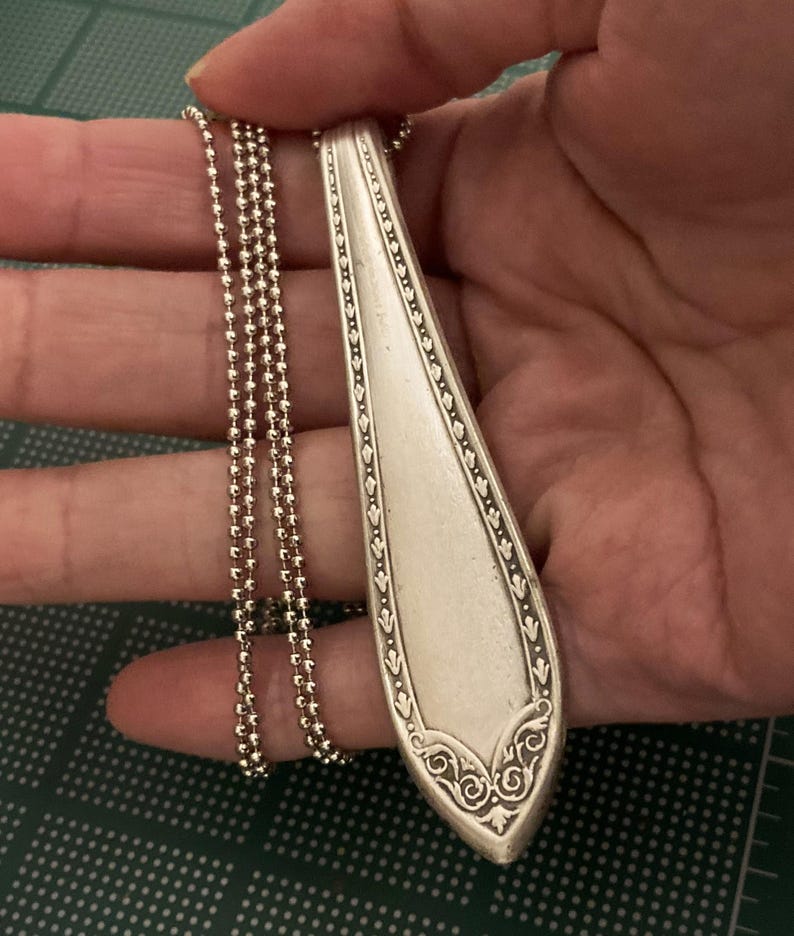 May include: A silver spoon pendant necklace with a decorative design on the handle. The necklace is made with a silver ball chain.