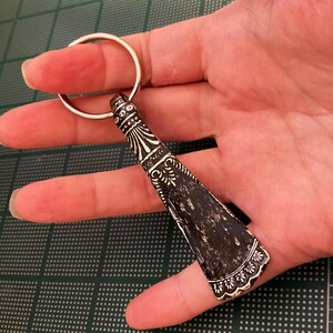 May include: A silver key chain with a decorative black and silver shoe horn charm. The shoe horn has an ornate design and a scalloped edge.
