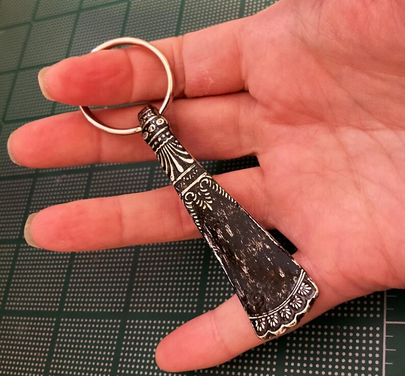 May include: A silver key chain with a decorative black and silver shoe horn charm. The shoe horn has an ornate design and a scalloped edge.