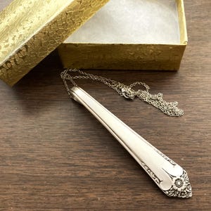 May include: Silver-toned necklace featuring a spoon-shaped pendant with floral detailing. The pendant is attached to a delicate chain, resting on a wooden surface next to a gold gift box.