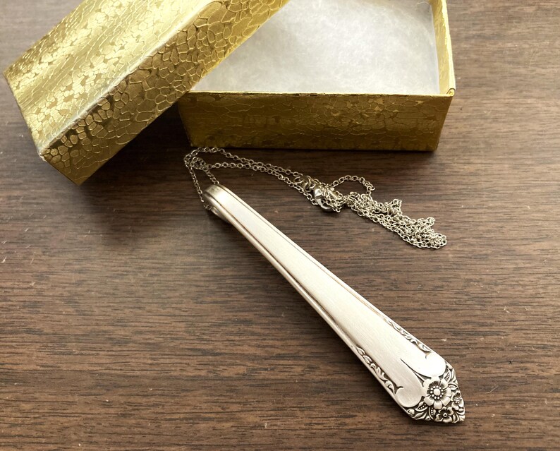 May include: Silver-toned necklace featuring a spoon-shaped pendant with floral detailing. The pendant is attached to a delicate chain, resting on a wooden surface next to a gold gift box.