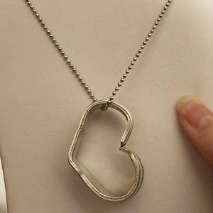 May include: A silver necklace with a heart-shaped pendant. The pendant is made of metal and has a slightly open design.