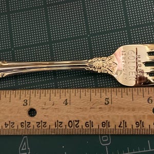 May include: A gold-colored fork with ornate detailing on the handle and the text "GOLDEN BIRTHDAY". The fork is placed next to a wooden ruler, showing measurements in inches. The fork is approximately 8 inches long.