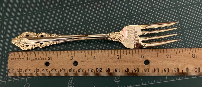 May include: A gold-colored fork with ornate detailing on the handle and the text "GOLDEN BIRTHDAY". The fork is placed next to a wooden ruler, showing measurements in inches. The fork is approximately 8 inches long.
