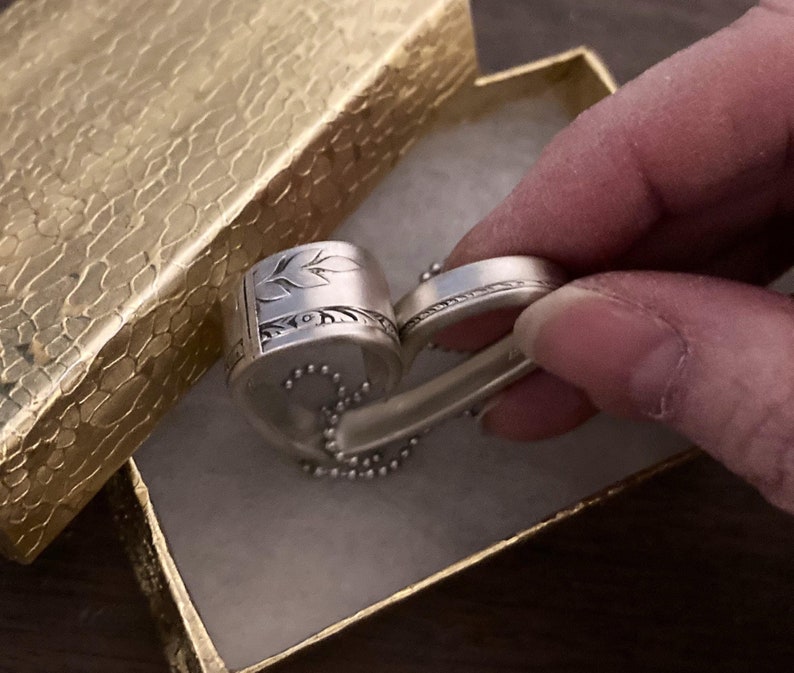 May include: A silver ring with a heart-shaped design and a floral pattern. The ring is in a small box with a gold-colored lining.