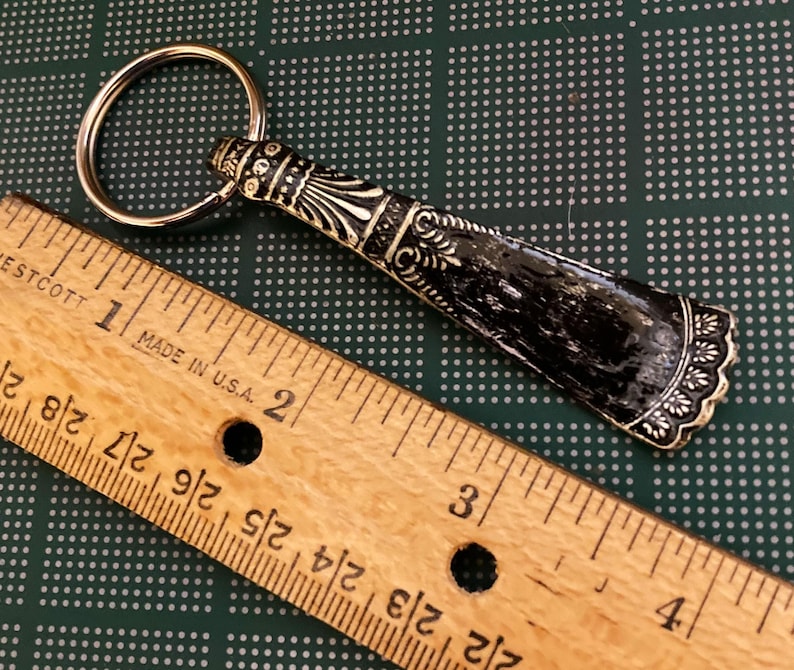 May include: A black and gold shoehorn keychain with a decorative design. The keychain is about 2 inches long and has a silver key ring.