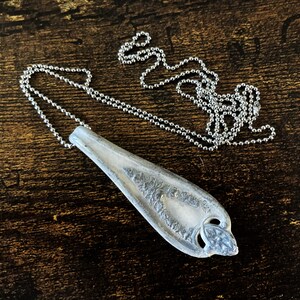 May include: A silver spoon pendant necklace with a detailed design on the spoon. The necklace is on a silver ball chain.