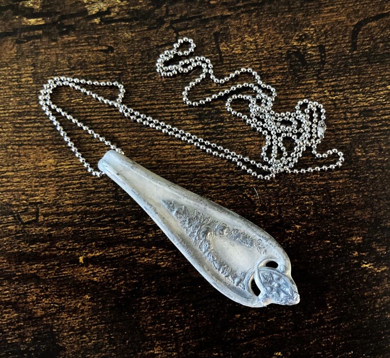 May include: A silver spoon pendant necklace with a detailed design on the spoon. The necklace is on a silver ball chain.