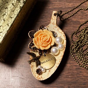 May include: A necklace made from a repurposed spoon with a peach-colored flower, a bird charm, and a round tag that says "BALANCE". The spoon is painted white with brown accents and has white pearl beads attached.